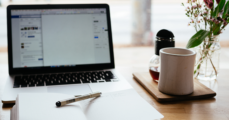 a computer, notepad and pen, and a mug on a table